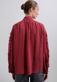 Mujer con el cabello recogido en un moño, viste una blusa roja de manga larga y holgada con detalles de volantes en las mangas y unos jeans gris oscuro, de espaldas.