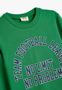 Zielona bluza z okrągłym dekoltem, z białym i granatowym napisem: "Drużyna Ligi Futbolowej Bez Ograniczeń, Bez Problemów". Materiał bawełniany.