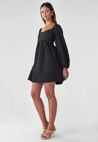 Schwarzes Kleid mit einem quadratischen Ausschnitt, strukturiertem Stoff und Puffärmeln. Kurzer Saum und leichte Raffung unter der Brust.