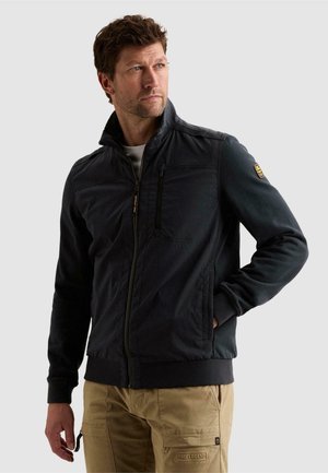 Homme tourné vers la gauche portant une veste noire zippée avec une poche poitrine et un pantalon cargo beige, main dans la poche, sur fond clair.