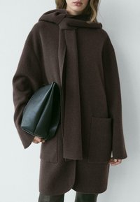 Manteau en laine marron avec capuche et détail d'écharpe, doté d'une poche avant et d'une coupe ample. Accompagné d'un sac en cuir noir.