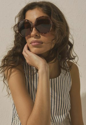 Moteris su banguotais rudais plaukais, dėvinti didelius apvalius akinius nuo saulės ir dryžuotą be rankovių palaidinę, atsirėmusi smakru į ranką priešais vienspalvį sieną.