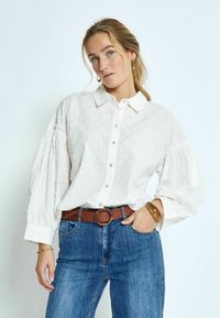 Witte blouse met een textuur van stippen, knoopsluiting en puffmouwen. Gepaard met een blauwe spijkerbroek en een leren bruine riem.