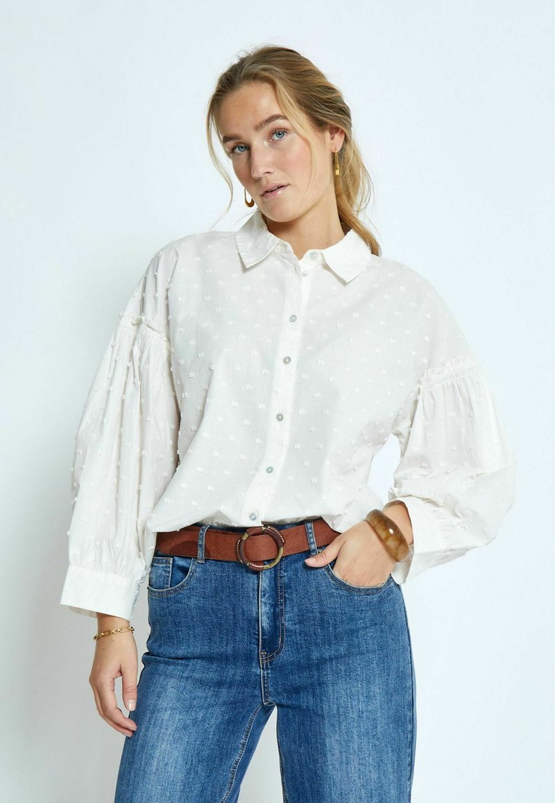 Witte blouse met een textuur van stippen, knoopsluiting en puffmouwen. Gepaard met een blauwe spijkerbroek en een leren bruine riem.