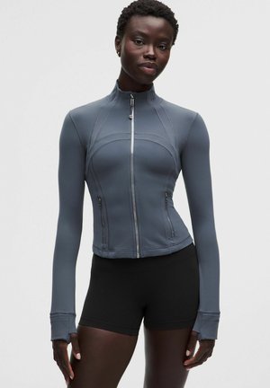 Frau trägt eine eng anliegende graue Jacke mit Reißverschluss und Daumenlöchern sowie schwarze Shorts, steht vor einem schlichten hellen Hintergrund.