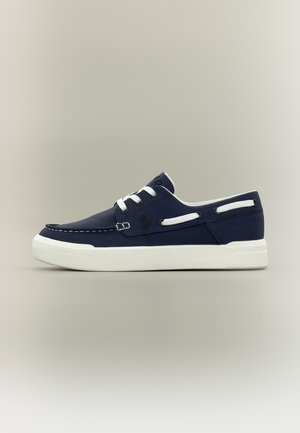 Baskets décontractées bleu marine avec semelle en caoutchouc blanche, lacets blancs et détails de lacets sur le côté, présentées sur un fond gris neutre.