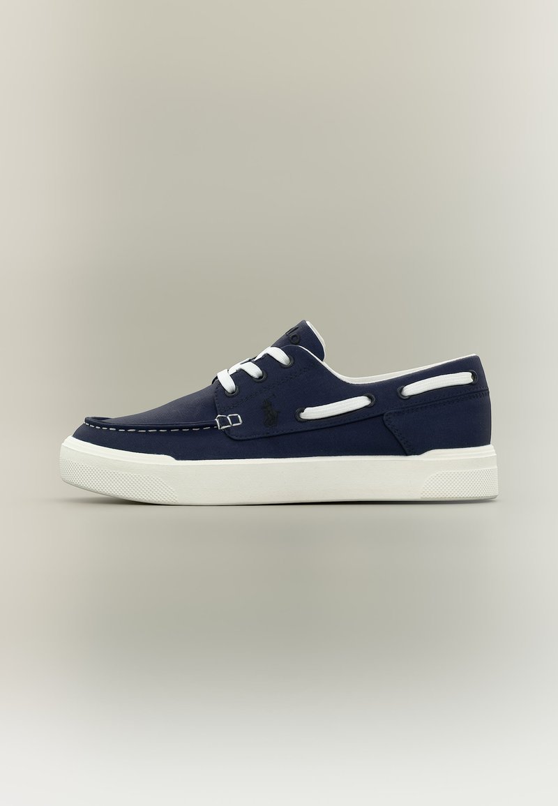 Καθημερινό αθλητικό παπούτσι σε ναυτικό μπλε με λευκή λαστιχένια σόλα, λευκά κορδόνια και λεπτομέρεια με κορδόνι στο πλάι, εμφανιζόμενο σε ουδέτερο γκρι φόντο.