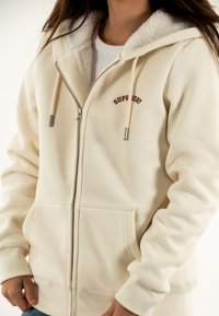 Sudadera con cremallera de color crema, con capucha forrada de suave sherpa, bordado que dice "Superdry", puños acanalados y dos bolsillos frontales.