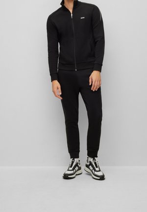 Mann trägt schwarze Jacke mit Reißverschluss und passende schwarze Jogginghosen, kombiniert mit weißen und schwarzen Turnschuhen vor einem schlichten grauen Hintergrund.