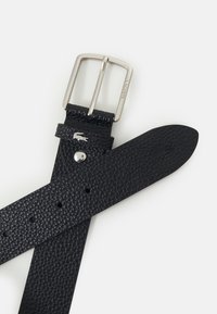 Ceinture en cuir noir avec une surface texturée, boucle en métal argenté et logo gravé. La sangle dispose de plusieurs trous pour l'ajustement.