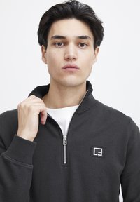 Zwarte zip-up sweatshirt met hoge kraag, voorzien van een klein logo-opdruk op de borst en geribde manchetten. Draagvlak over een wit T-shirt.