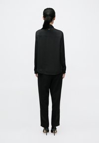 Čierna saténová blúzka s dlhými rukávmi a zaobleným lemom, v kombinácii s čiernymi zúženými nohavicami. Na zadnej strane sa pri golieri nachádza jemné logo.