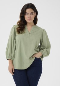 Ljusgrön blus med vertikala ränder, V-hals och puffärmar. Mjuk kvalitet, avslappnad passform och elastiska mudd. Bärs med mörkblå jeans.