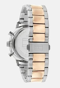 Metalarmband med länk i silver- och guldtonad rostfritt stål. Rund boett med tryckknappar och en minimalistisk vit urtavla.