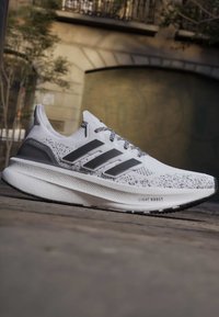 Chaussure de course Adidas Ultraboost blanche et noire avec tige en maille, trois bandes sur le côté, et semelle Light Boost sur pavé extérieur.