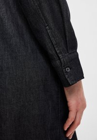 Σκούρο τζιν πουκάμισο με κουμπωτό μανίκι, λείο ύφασμα και στενή εφαρμογή. Ορατή ραφή κατά μήκος τ�ου χεριού και της περιοχής του καρπού.