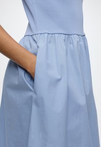 Personne portant une robe sans manches bleu clair, montrée de la taille au genou, avec une main dans une poche latérale.