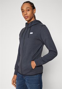 Tamno siva zip-up dukserica s kapuljačom, koja ima vezice, prednji džep i mali logo na prsima. Mekana, teksturirana tkanina.
