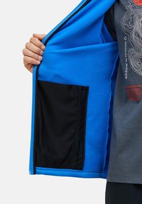 Giacca in pile azzurro chiaro con tasca interna nera, dotata di zip e texture morbida, accompagnata da una t-shirt grigia con stampa sottostante.