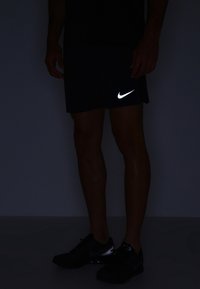 Pantalones cortos negros de Nike con un logo reflectante, fabricados con material ligero. Exhibidos en condiciones de poca luz, resaltando el brillante contraste del logo.