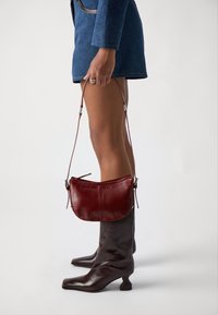 Borsa in pelle rossa di forma arrotondata, con una sola tracolla e dettagli di cucitura a vista, tenuta da una figura che indossa stivali marroni fino al ginocchio.