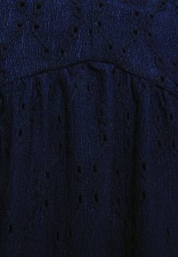 Tecido texturizado azul marinho com padrão elevado e pequenos detalhes recortados; a cintura franzida cria uma forma suave e fluida.