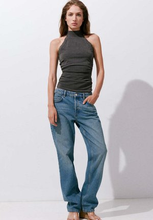 Femme portant un top dos nu gris foncé et un jean bleu ample, debout avec une main dans la poche sur un fond blanc uni.