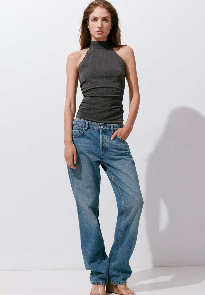Femme portant un top dos nu gris foncé et un jean bleu ample, debout avec une main dans la poche sur un fond blanc uni.