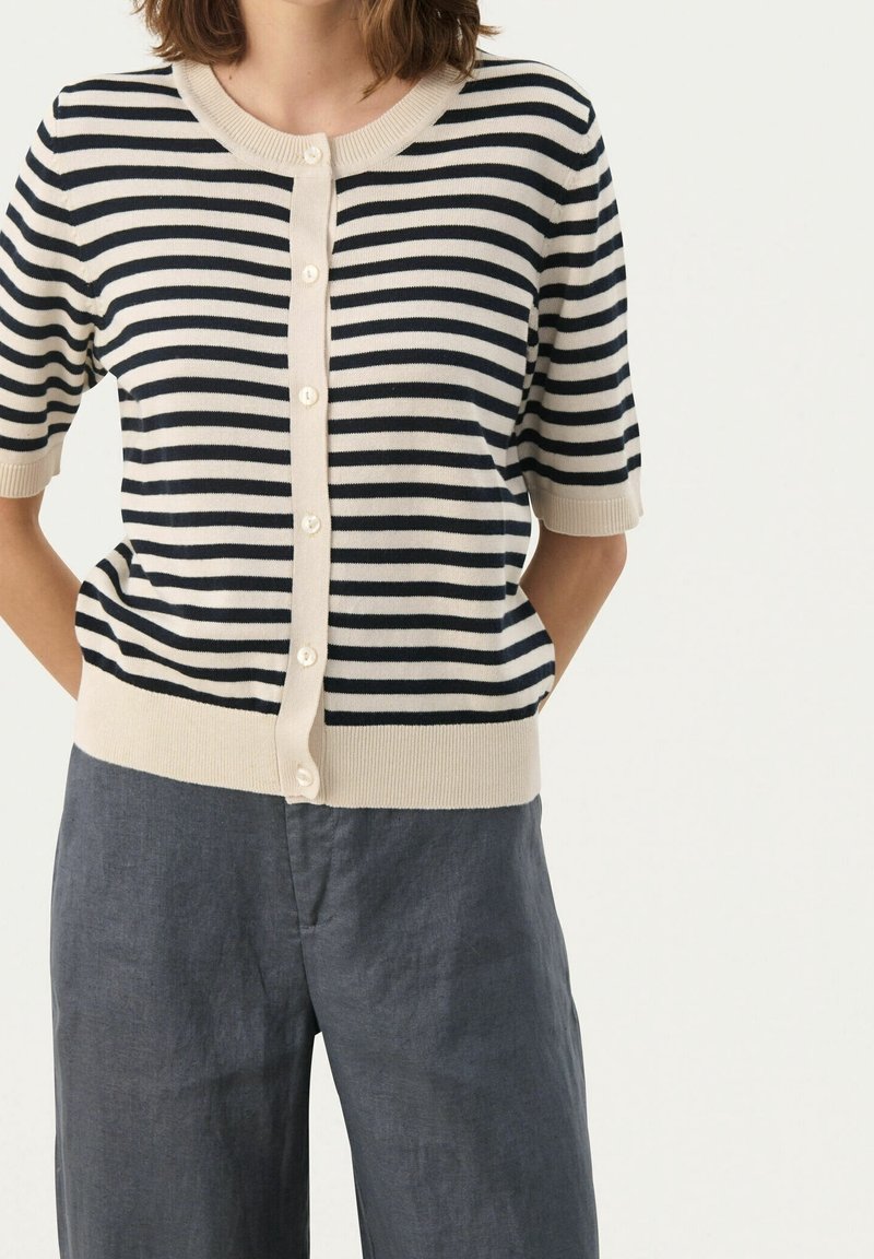 Femme portant un cardigan à manches courtes, boutonné, rayé noir et crème, avec un pantalon gris taille haute, mains dans le dos.