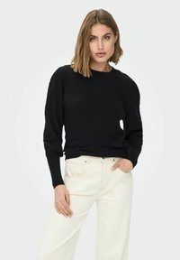 Svart strikket genser med en teksturert, diamantmønster, ribbestrikkede mansjetter og rund hals, kombinert med hvite vide jeans.