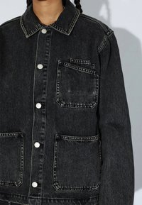 Veste en denim noire avec fermeture à boutons et trois poches plaquées sur le devant, portée par une personne aux cheveux tressés.