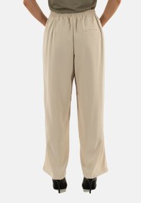 Pantalons beige légers, à taille élastique, avec des jambes larges et une poche arrière, associés à des chaussures à talons noirs. Texture lisse.