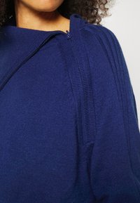 Maglione blu navy a collo alto, con dettagli a zip e accenti a costine sulle spalle, che mette in mostra una texture morbida.