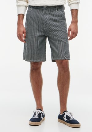Mann trägt schwarz-weiß vertikal gestreifte Shorts mit marineblauen Canvas-Schuhen und weißem Pullover, steht vor weißem Hintergrund.