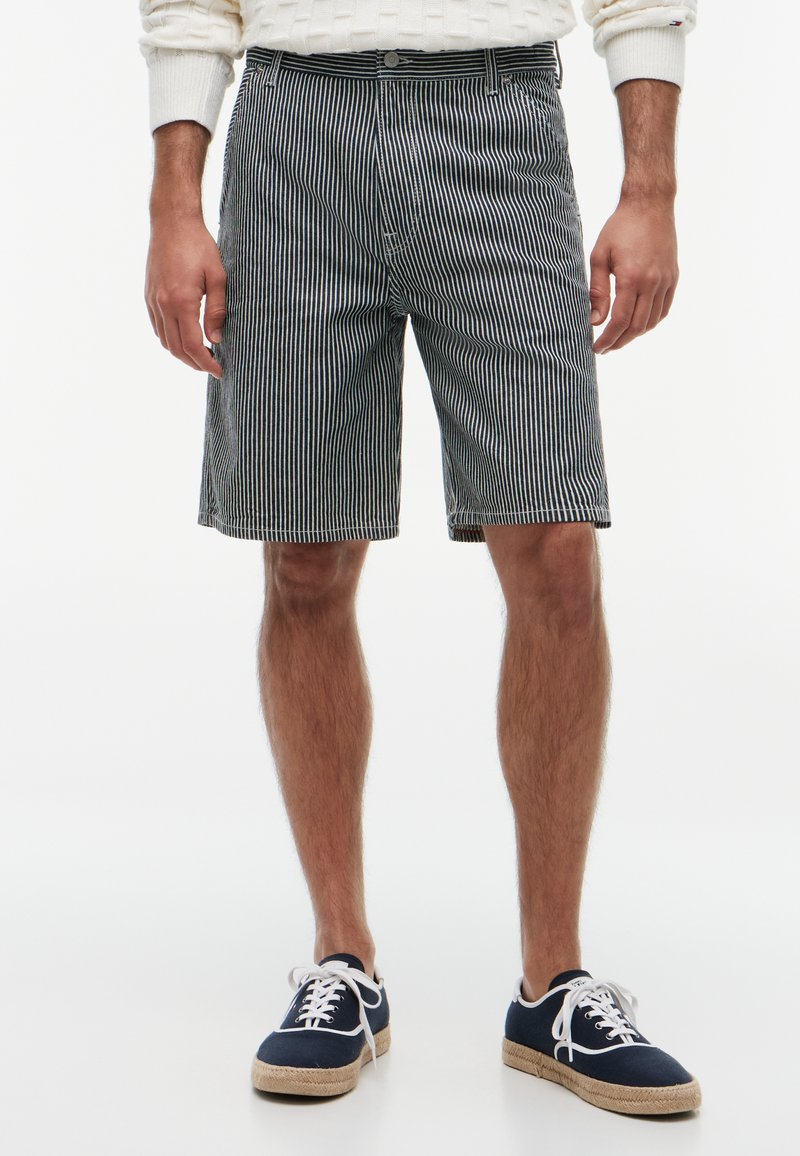 Homme portant un short rayé vertical noir et blanc, des chaussures en toile bleu marine et un pull blanc, debout sur fond blanc.