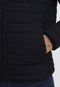 Črna puffer jakna s horizontalnim prešitjem, ki ima stransko žep in mehko, gladko teksturo. Je lahka in oprijeta.