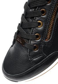 Close-up van een zwarte leren sneaker met getextureerde veters, bronzen veteroogjes en een gouden rits aan de zijkant met gedetailleerd stiksel.