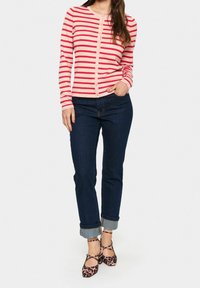 Roter und pink gestreifter Cardigan mit Knöpfen, getragen über dunklen Jeans, kombiniert mit Leopardenmuster-Ballerinas. Glatter Stoff, tailliertes Design, lange Ärmel.