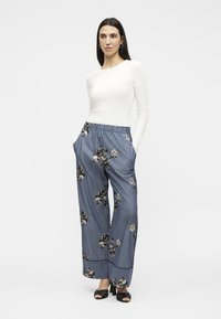 Femme aux longs cheveux foncés portant un haut blanc à manches longues, un pantalon large à rayures bleues avec imprimé floral et des talons ouverts noirs.