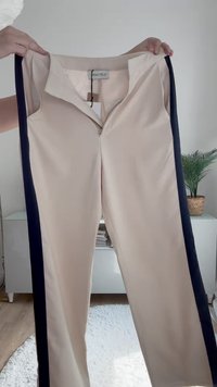 Pantalones beige con franjas laterales en azul marino oscuro, cierre con cremallera en la parte frontal y cuello. El material parece suave; etiqueta visible en el interior.