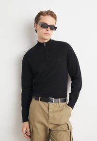 Pulover tricotat negru cu guler înalt și închidere cu fermoar, purtat cu pantaloni cargo bej și curea neagră. Accesoriile includ ochelari de soare pătrați.