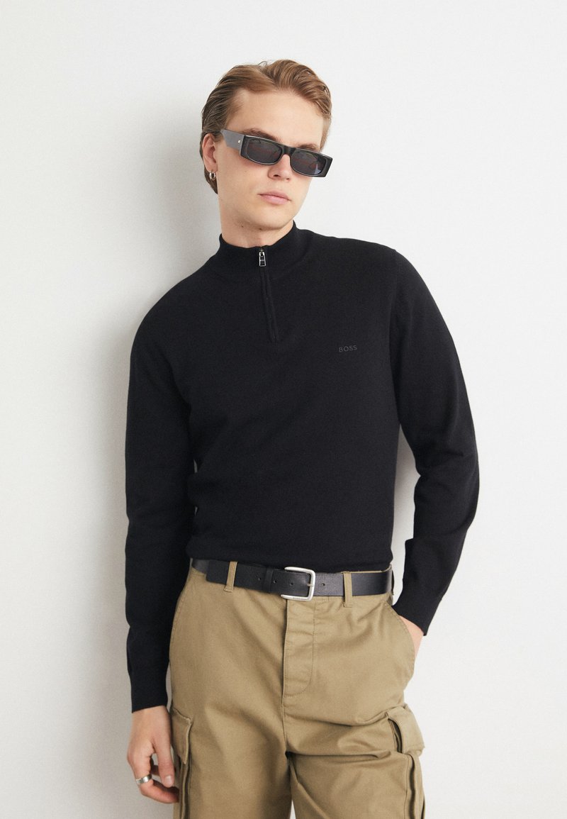 Pulover tricotat negru cu guler înalt și închidere cu fermoar, purtat cu pantaloni cargo bej și curea neagră. Accesoriile includ ochelari de soare pătrați.