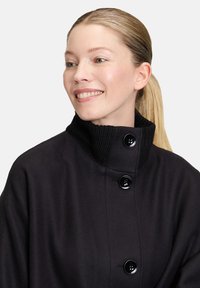 Czarny wełniany płaszcz z prążkowanym kołnierzem, posiadający trzy duże czarne guziki oraz gładką fakturę. Zaprojektowany z myślą o dopasowanym kroju.