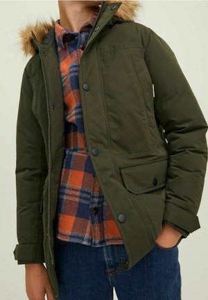 Personne portant une parka vert olive avec capuche en fourrure, par-dessus une chemise à carreaux orange et bleue et un jean bleu, debout avec une main dans la poche.