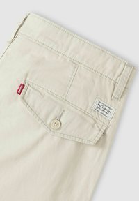 Beige katoenen broek met een achterzak voorzien van een knoop en een merklabel. De stof heeft een gladde textuur en eenvoudige stikseldetails.