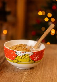 Ciotola di ceramica con un personaggio verde sorridente, design rosso e verde con fiocchi di neve, riempita di cereali e latte, cucchiaio di legno all'interno.