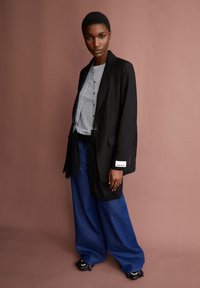 Vrouw met kort haar, gekleed in een zwarte blazer, grijze knopenoverhemden, blauwe wijde broek en zwarte schoenen met franjes, staande tegen een bruine muur.