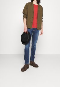 Oliivinvihreä takki nappien kanssa, punainen t-paita, siniset farkut, ruskeat saappaat ja musta hattu kädessä. Sujuvat tekstuurit ja rento muotoilu.