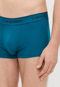 Zaļgani zili apakšbiksītes izstiepjama materiāla ar melnu jostas joslu, kurā atrodas Calvin Klein logotips. Tām ir pieguļošs dizains un gluds tekstūras izskats.