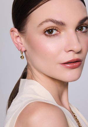 Złote kolczyki w formie kół z okrągłymi zawieszkami, gładka tekstura i błyszczące wykończenie. Lekka tkanina opadająca na ramię, miękki neutralny odcień.